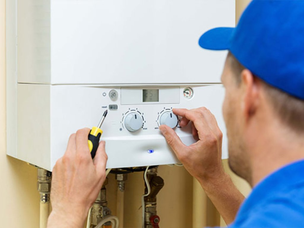 A worker wearing a blue cap adjusts the controls of a wall-mounted boiler with one hand while holding a screwdriver in the other hand.