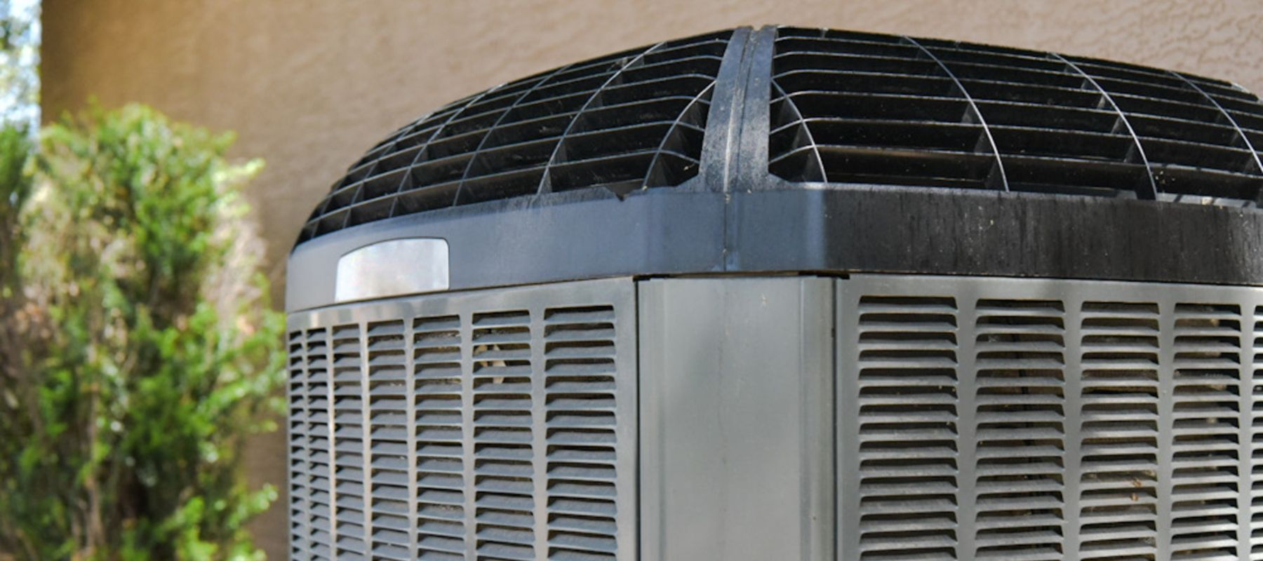 A close-up view of a residential outdoor air conditioning unit with a metal grille and black vented top, positioned next to a beige wall and green shrubbery.