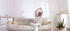 A woman in white pajamas sits on a beige sofa in a bright, modern living room, stretching her arms above her head and smiling. Sunlight streams in through window blinds, casting shadows on the wall.
